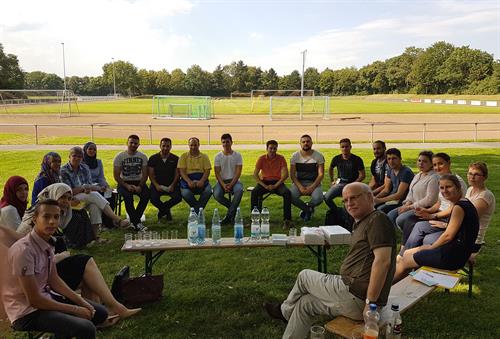 Auf einer Wiese sitzen in einem Kreis etwa 20 Personen - zumeist junge Männer und Frauen auf Bänken. In der Mitte steht eine Bank mit Wasserflaschen und Gläsern darauf. Im Hintergrund sieht man einen Sportplatz mit Toren und blauen Himmel.